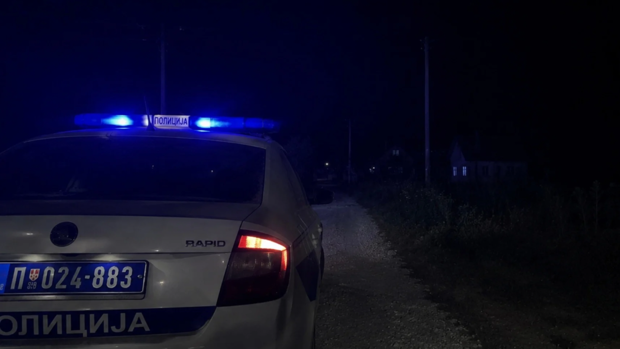Policijsko vozilo sa uključenim rotacionim svetlima na seoskom putu tokom noći u selu &Scaron;krijelje kod Tutina. U pozadini se naziru kuće i mračni ruralni pejzaž, dok plava svetla policije osvetljavaju okolinu nakon tragičnog događaja u kojem je sedamnaestogodi&scaron;njak izgubio život prilikom či&scaron;ćenja lovačke pu&scaron;ke.